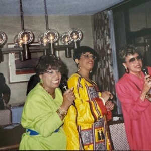 Three women singing with microphones in front of a table.