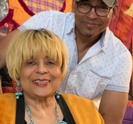 A man and woman posing for a photo in front of a colorful quilt.